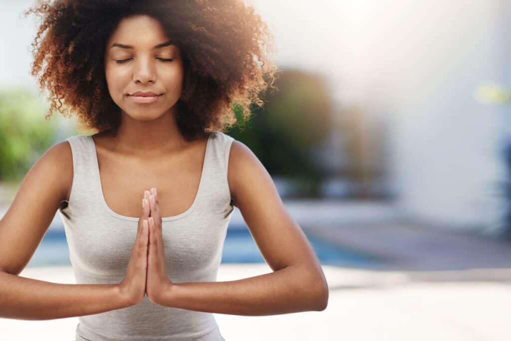 With closed eyes and curly hair, the person stands outdoors in a peaceful pose, hands in prayer position—a serene moment embodying self-care activities—while wearing a gray sleeveless top.