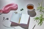 A pink sleep mask, open notebook with a pen, earphones, two capsules, a cup of tea, and a small plant sit on a light-colored surface, creating the perfect setup for evening self-care routines.