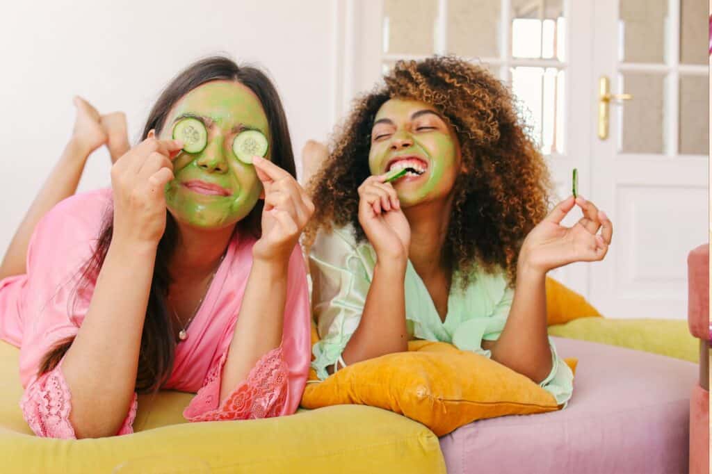 Two women with face masks and cucumbers, smiling and relaxing on colorful cushions, indulge in their evening self-care routines indoors.