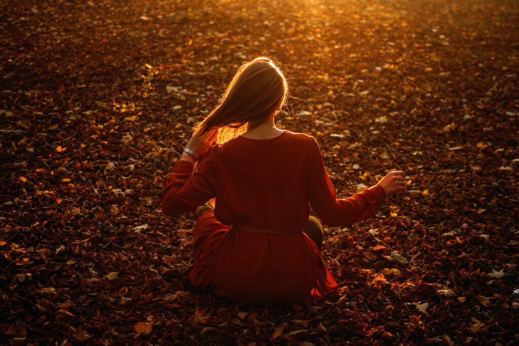 Person sitting on fallen leaves in a forest, with long hair illuminated by a warm, golden sunset, embracing evening self-care routines amidst nature's tranquil embrace.