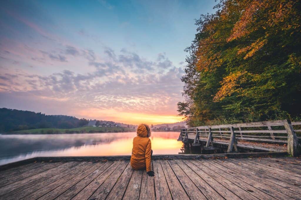 A person in a brown jacket sits on a wooden dock, embracing evening self-care routines while facing a lake surrounded by trees with autumn foliage, under a cloudy sky at sunset.