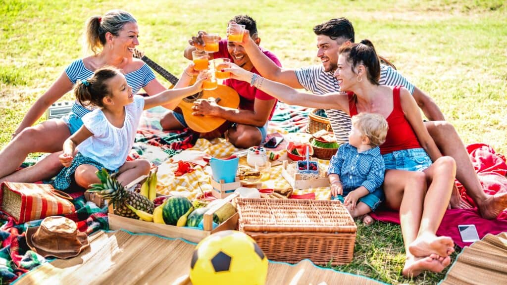 A group of adults and children sit on blankets in a park, sharing a picnic and raising drinks together—a perfect example of frugal living tips, with homemade food and outdoor fun instead of expensive outings.