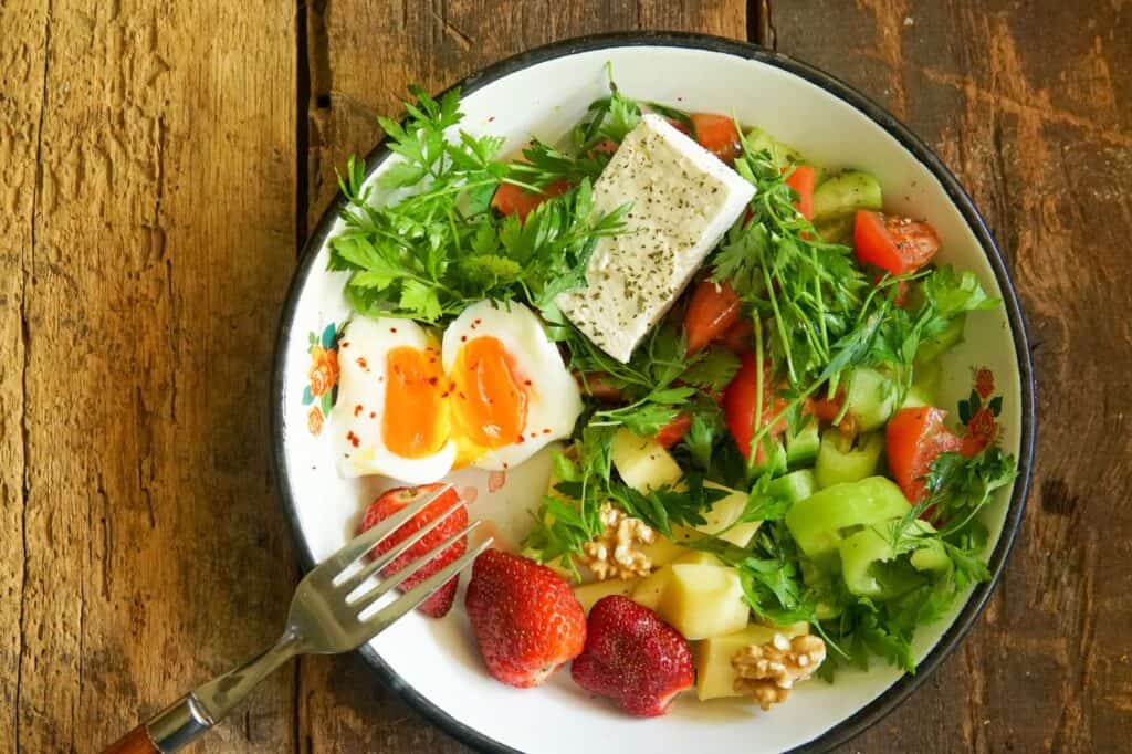 A plate of food featuring soft-boiled eggs, strawberries, walnuts, cheese, and mixed vegetables rests on a wooden table, inviting mindful self-care with every bite.