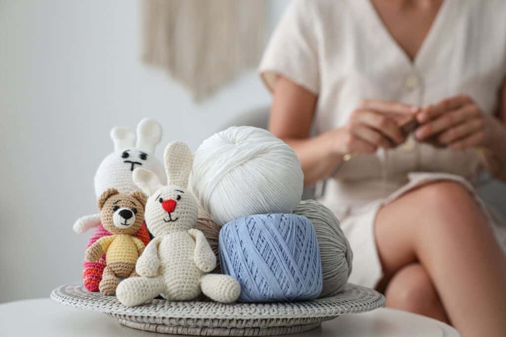 A person sits knitting in mindful self-care beside a basket containing balls of yarn and knitted toy animals, including rabbits and a bear, on a woven surface.