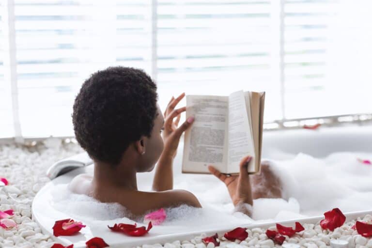 Enjoying a moment of mindful self-care, the person relaxes in a bubble bath, immersed in a book. Surrounded by rose petals and white pebbles, with soft light filtering through the blinds, this serene setting invites tranquility and reflection.