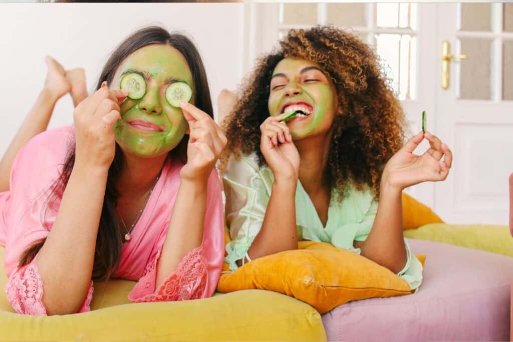 Two women, indulging in self-care activities, lie on cushions with green face masks. One holds cucumber slices over her eyes, both smiling and relaxed.