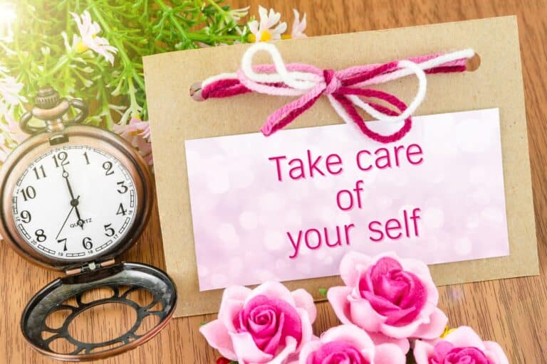 A pocket watch, flowers, and a card tied with pink yarn display the message "Take care of yourself" on a wooden surface.