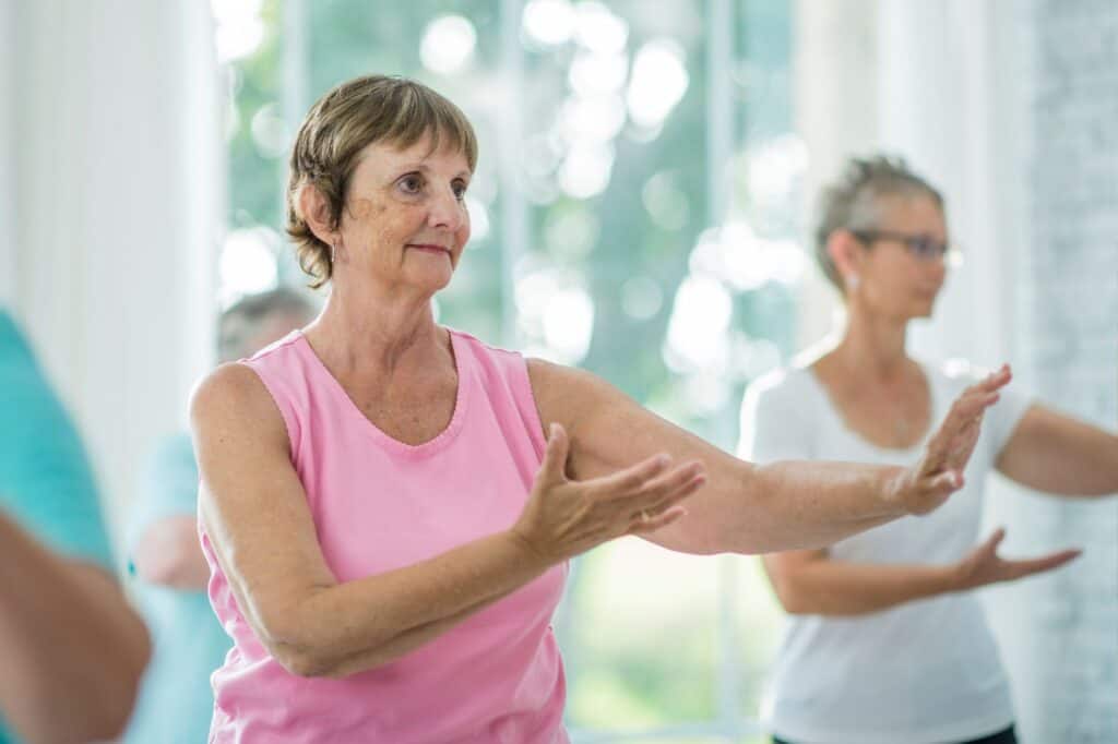 Three older women engage in a tai chi class, moving their arms with focus and concentration as part of their morning self-care rituals.