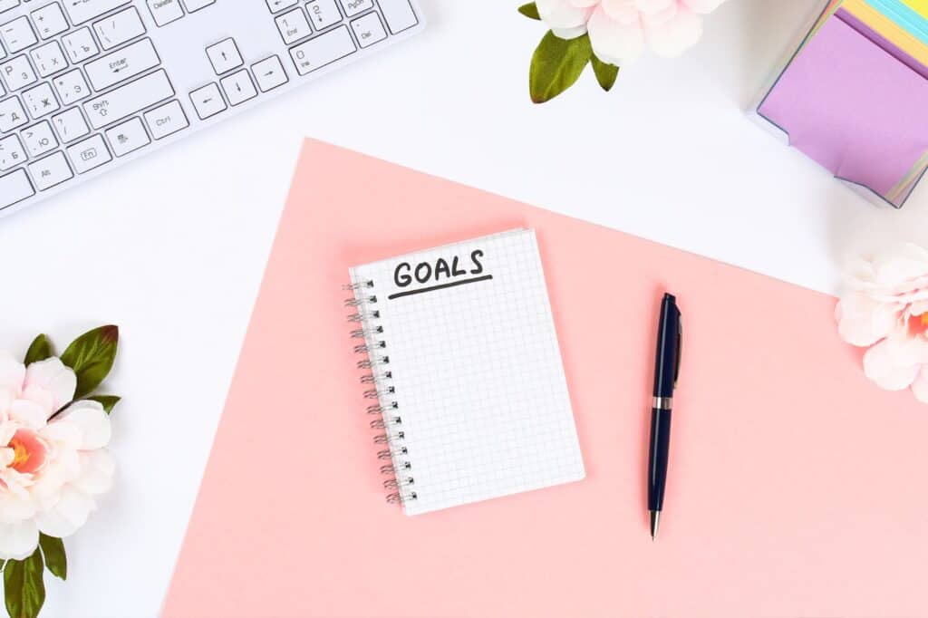 A notepad labeled "Goals" is placed on a pink paper with a pen, surrounded by flowers and next to a keyboard, offering inspiration for morning self-care rituals.