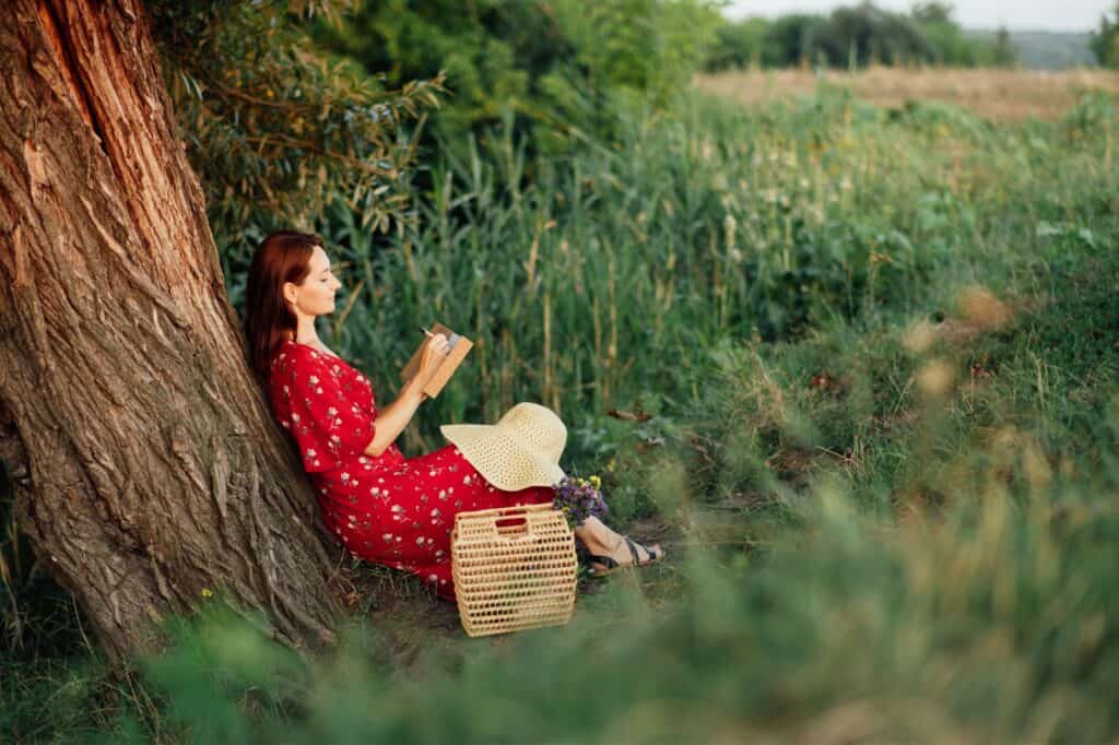 Woman in a red dress sitting against a tree, reading a book in a grassy field. A straw hat and woven bag with flowers are beside her.