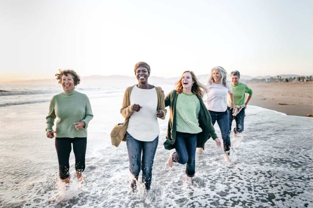 Five people laughing and walking through the surf on a beach at sunset, wearing casual clothing.