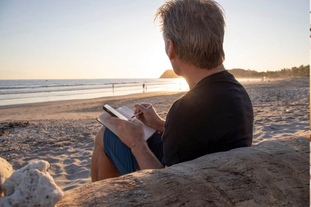 As the sun sets, a person sits on a log at the beach, writing in a notebook. The gentle rhythm of the ocean and distant figures create a serene backdrop, embodying evening tranquility—a perfect counterbalance to morning self-care rituals.