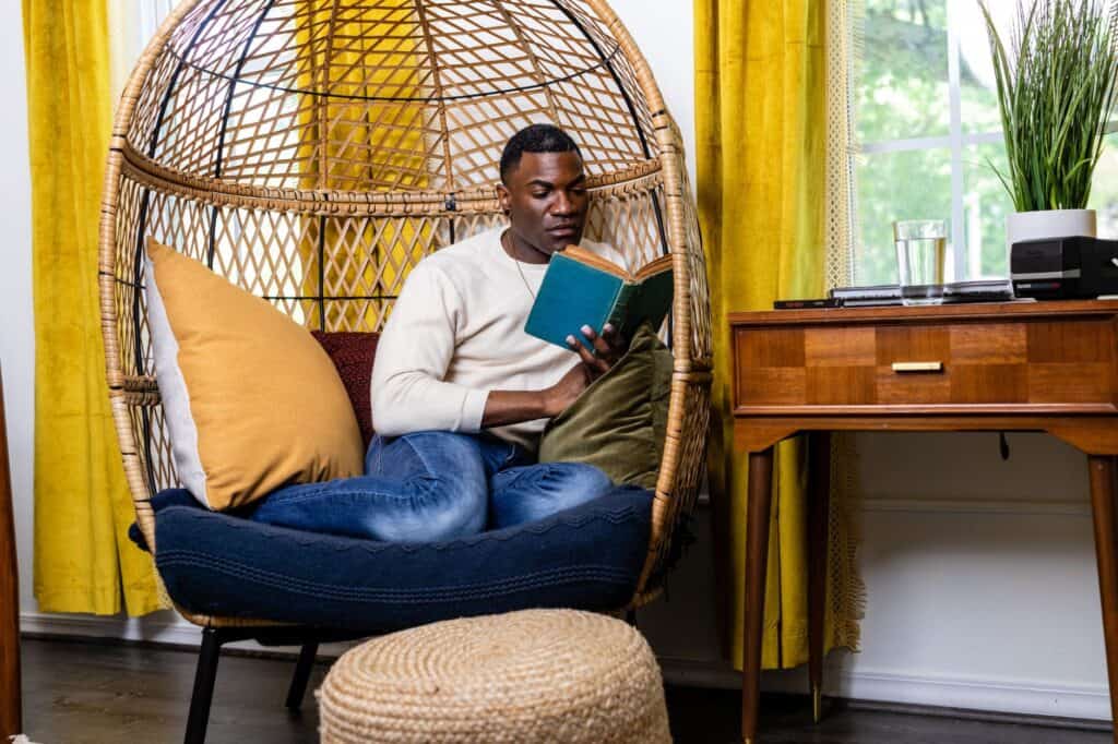 A person sits in a wicker chair, indulging in morning self-care rituals by reading a book. The room is accentuated with yellow curtains, a wooden desk, and a round ottoman.