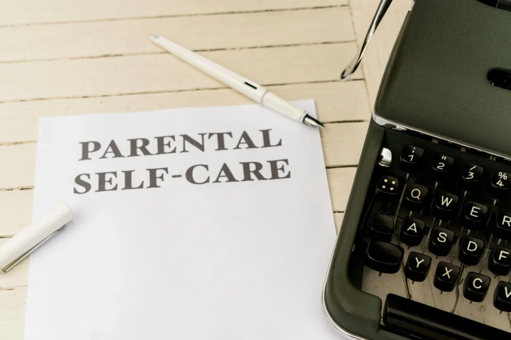 A paper reading "Parental Self-Care" is placed next to a typewriter and pen on a rustic wooden surface, emphasizing the timeless importance of self-care.