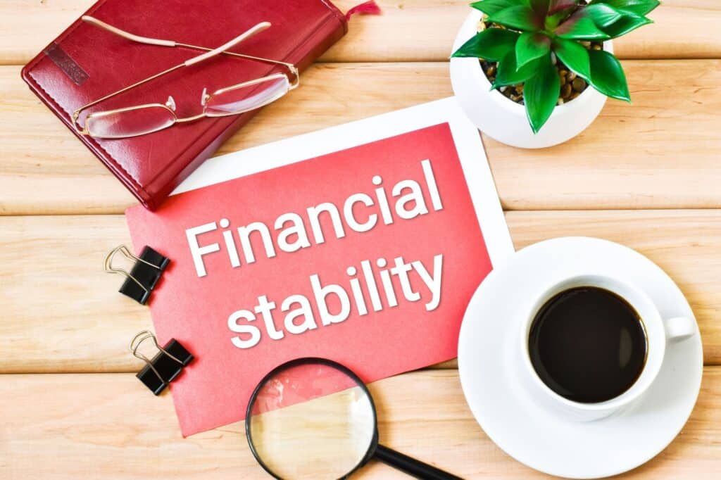 A document labeled "Financial stability" sits on a wooden desk alongside a steaming coffee cup, a magnifying glass, glasses, a vibrant green plant, and a striking red notebook, creating an atmosphere of focus and purpose.