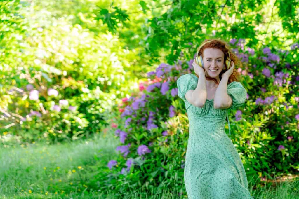 A woman in a green dress exudes confidence as she smiles, wearing headphones amidst a garden bursting with colorful flowers and lush greenery.