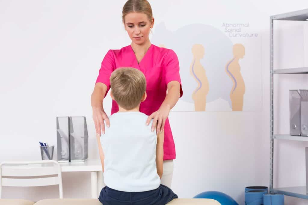 A healthcare professional examines a young boy's back in a medical office, exuding confidence with each precise movement. A poster about spinal curvature is visible in the background, underscoring the importance of expertise and assurance in medical care.