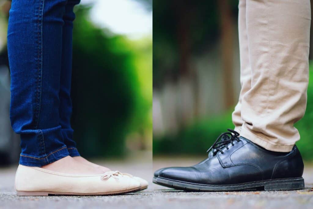 Two people stand face-to-face, depicted from the knees down. One wears jeans and beige flats, effortlessly exuding confidence, while the other sports light pants and black shoes. The backdrop of blurred greenery adds a touch of calm to this poised encounter.