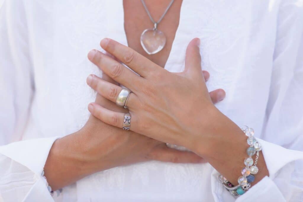 With rings and bracelets adorning their hands resting on a white garment, this person subtly exudes confidence; the heart-shaped pendant shines brightly as if echoing their inner strength.