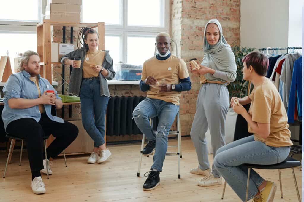 A diverse group of five people exudes confidence as they chat casually in a room, holding takeout containers. Some are seated while others stand near garments on a rack, each effortlessly showcasing how to exude confidence through their relaxed demeanor and engaging conversation.