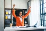 A person in an orange blazer sits at a desk with a laptop, arms raised in celebration, showcasing the joy of practicing self-care tips for busy professionals. A cup of coffee and neatly organized documents complete this moment of triumph and balance.