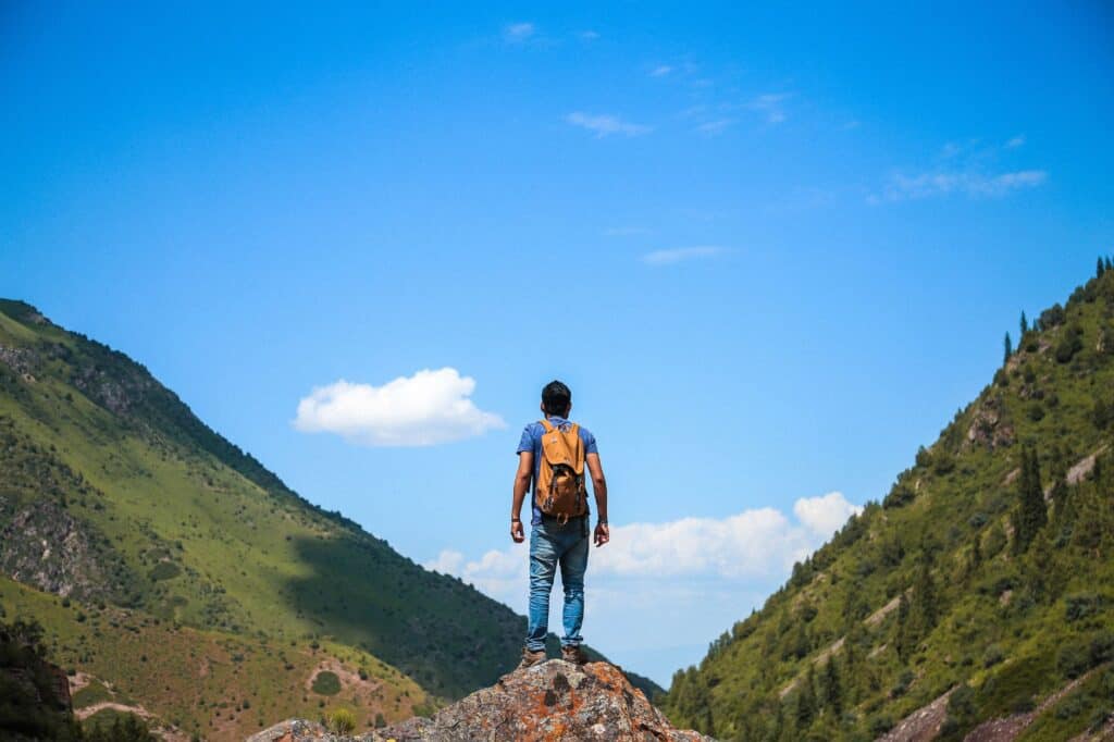 A person with a backpack stands on a rocky outcrop, gazing at the mountainous landscape framed by a vibrant blue sky, perhaps contemplating journal prompts that inspire their next great adventure.