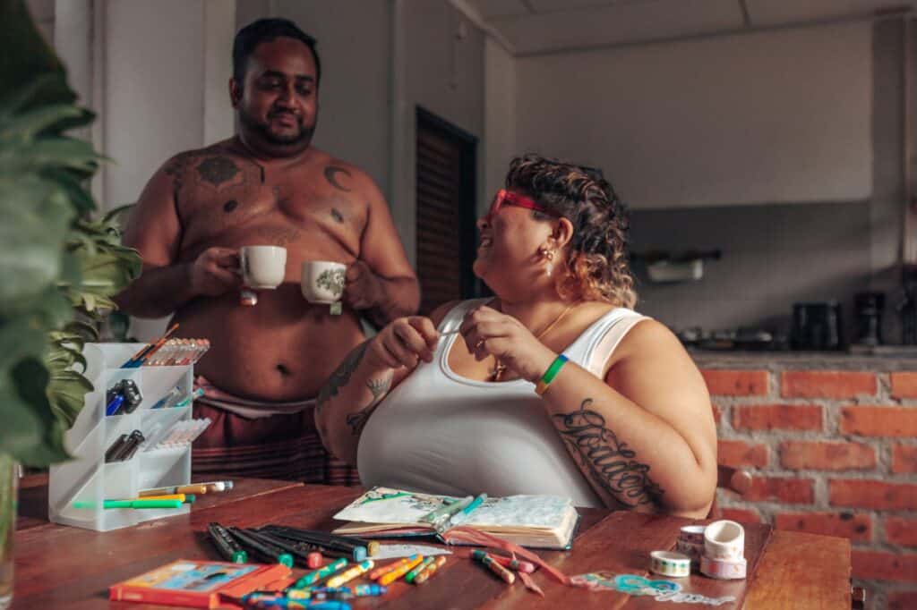 Two people in a cozy kitchen: one stands shirtless, holding mugs, while the other, wearing red glasses, is seated with stationery and an open notebook filled with journal prompts.