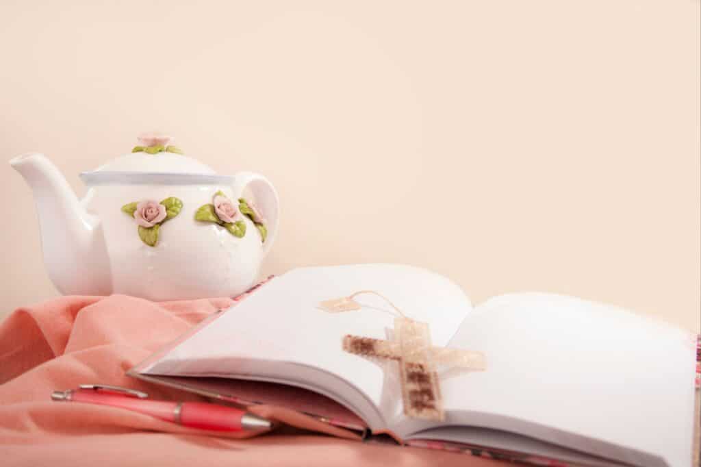 An open book with a woven bookmark lies invitingly next to a pink pen, ready for your journal prompts. A floral teapot adds a touch of charm on the soft pink fabric backdrop, setting the scene for inspired thoughts and reflections.