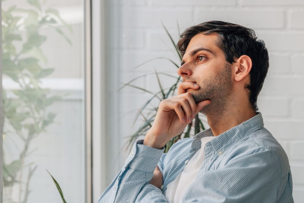 A man in a striped shirt sits pensively by the window, surrounded by lush plants, as if contemplating his next journal prompts.