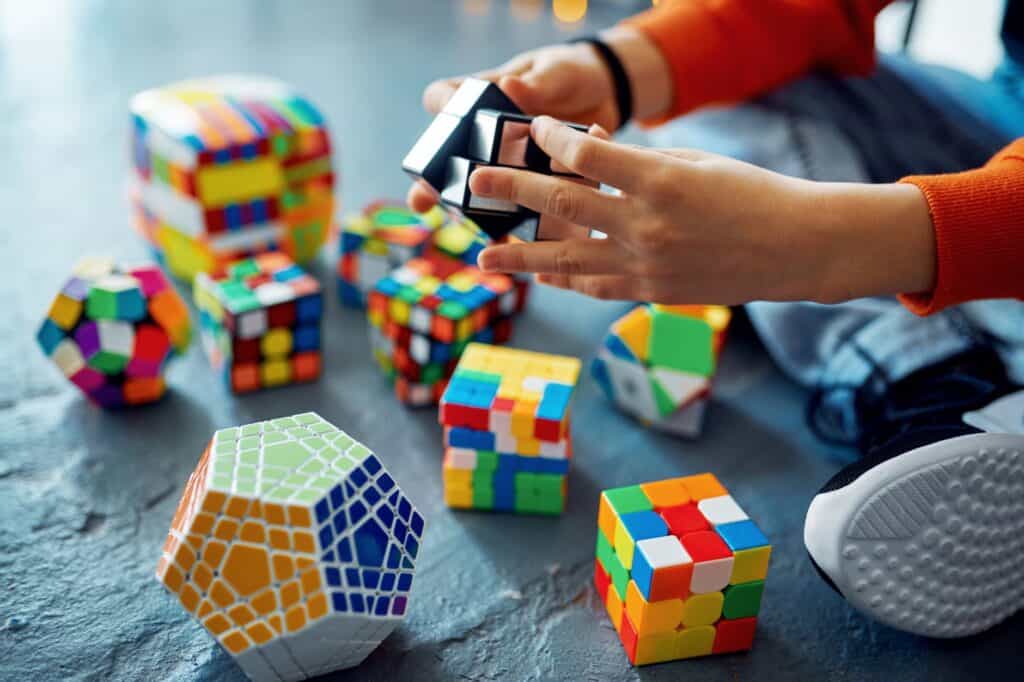 Amid various colorful Rubik's cubes and geometric puzzles on the floor, a person holds a puzzle cube, deep in thought as if searching for the perfect journal prompt hidden within its twisting pieces.