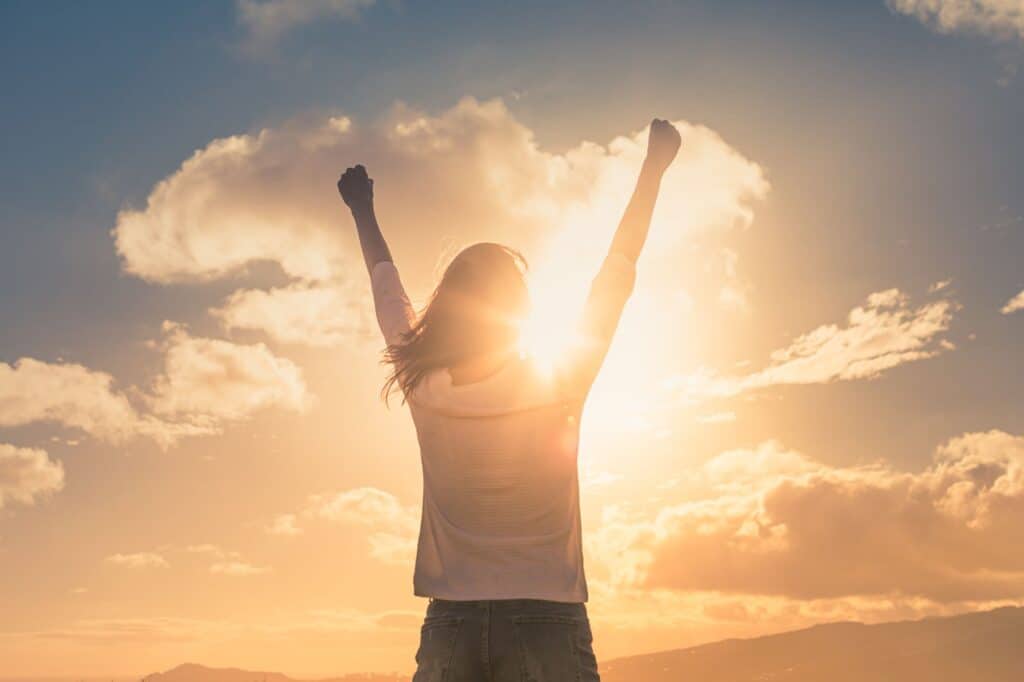 A person stands with raised arms, silhouetted against a bright sunset sky with scattered clouds, reminiscent of the inspiration found in journal prompts.