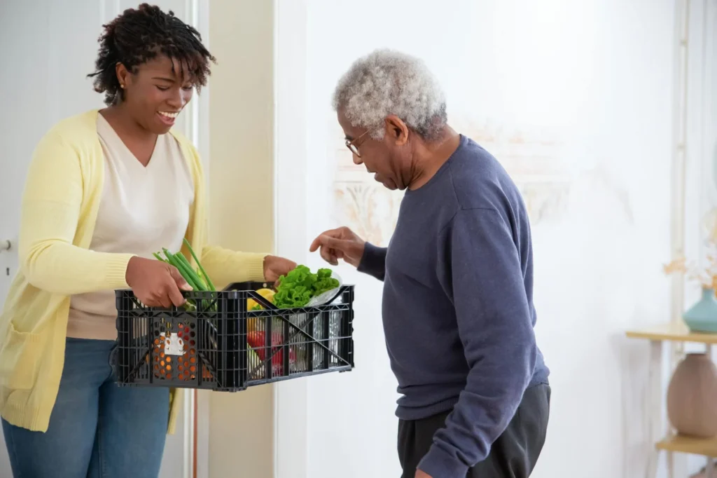 A woman in a yellow cardigan, embracing self-care activities through community connection, hands a crate of fresh vegetables to an elderly man in a blue sweater.