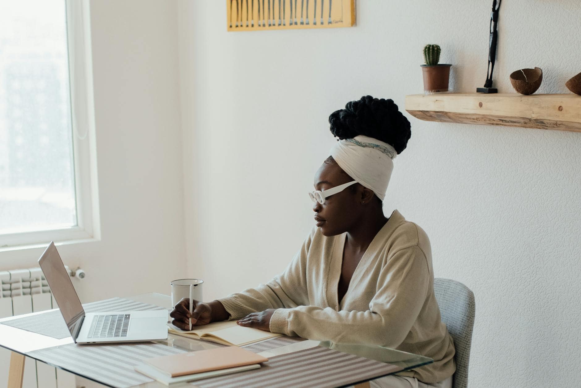 African American woman writing in a journal while working from home with a laptop.