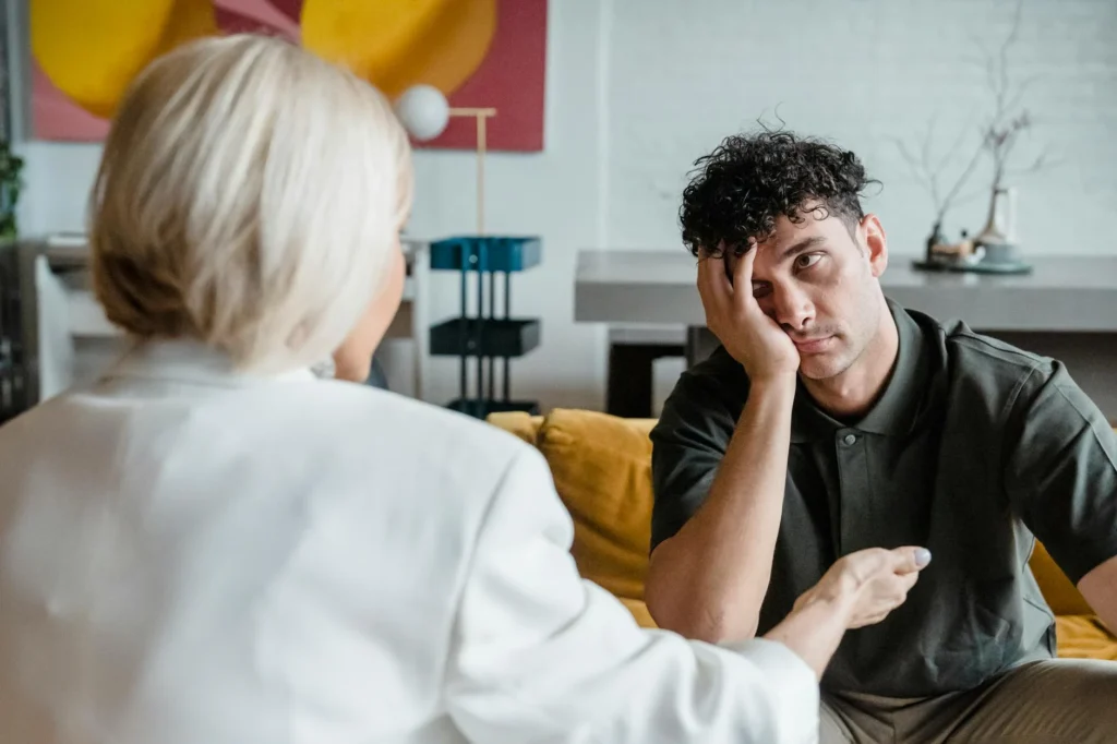 A man sitting on a yellow couch looks frustrated, his head resting on his hand, contemplating what to do when you feel lost. He's talking to a person with blonde hair in a white shirt, seeking guidance in the modern living room setting.