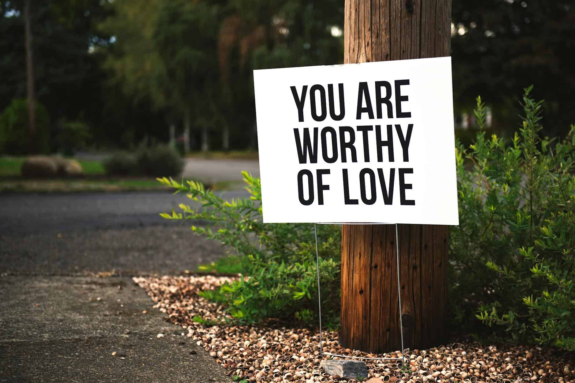 A positive message sign "You are worthy of love" in a park setting.