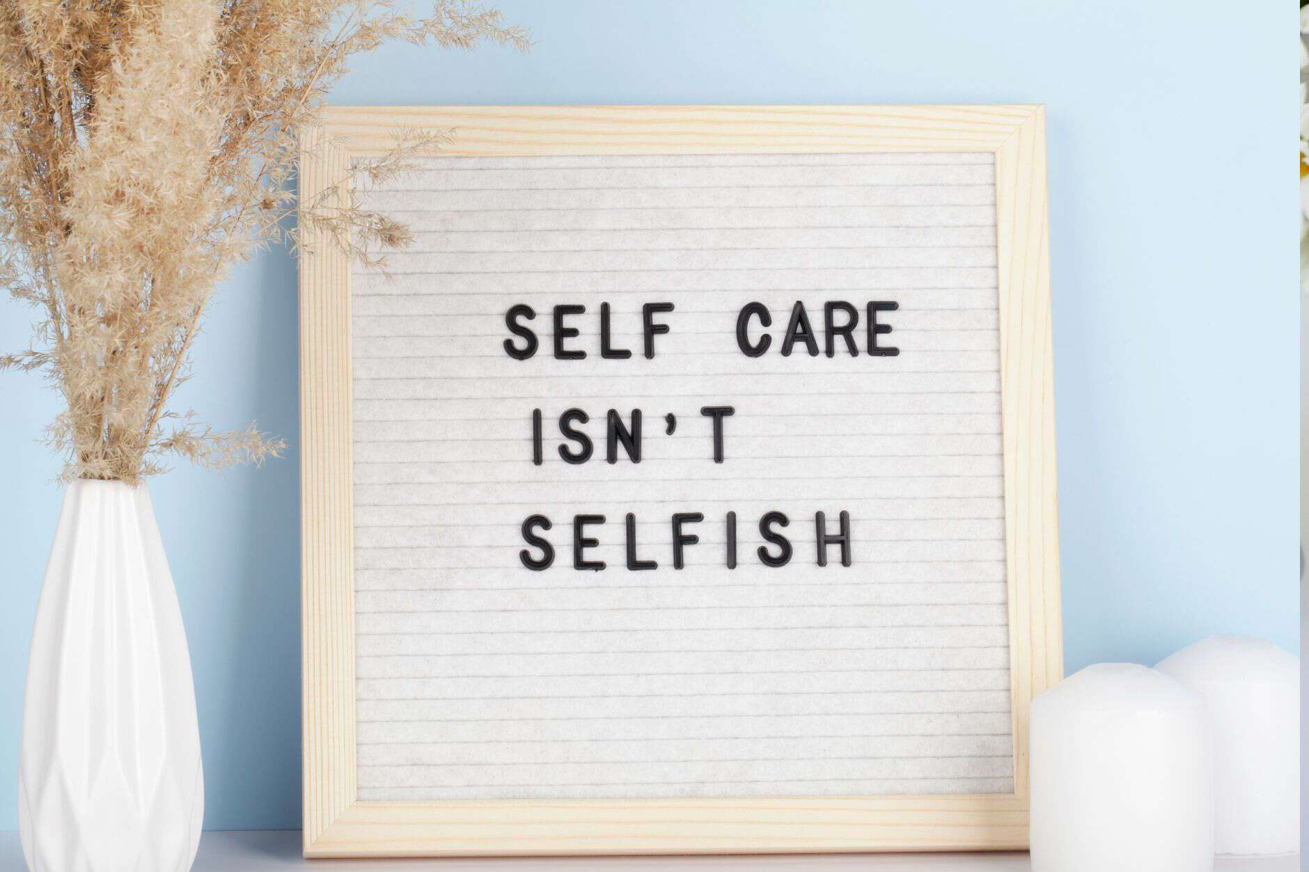 A letter board with the message "Self care isn't selfish" is displayed beside a white vase with dried flowers and two white candles, set against a light blue background. This tranquil scene invites contemplation of professional self-care ideas.