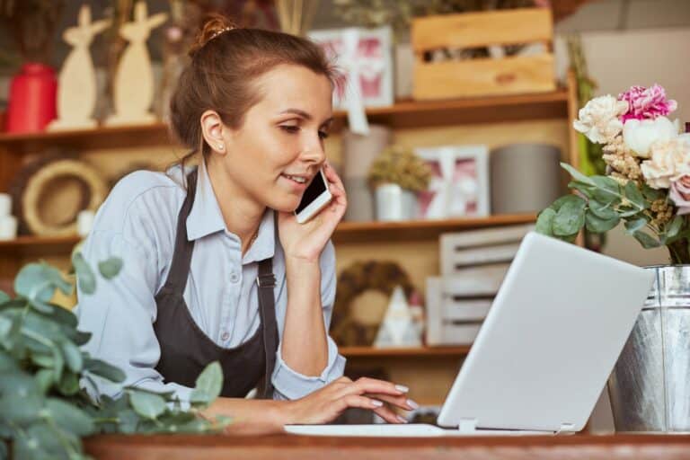 A florist, devoted to her craft and professional self-care ideas, wears an apron as she multitasks in the flower shop, discussing orders on the phone while deftly managing her laptop.