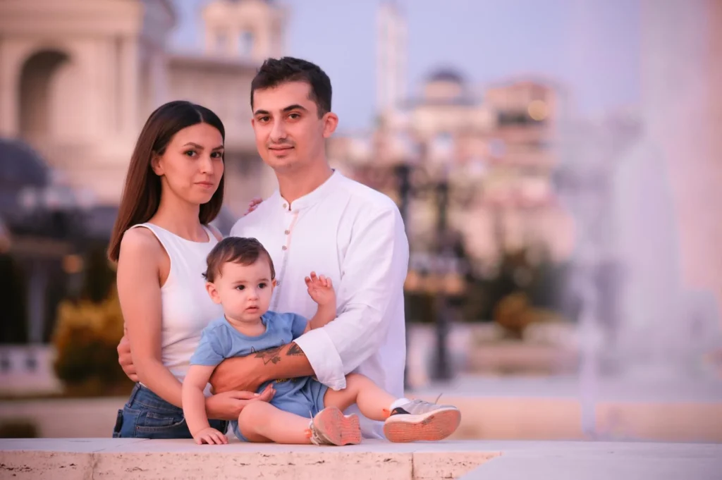 A loving family of three poses outdoors; a woman and a man hold a toddler, finding joy in the simple moment. The background features a blurred cityscape with classical architecture.