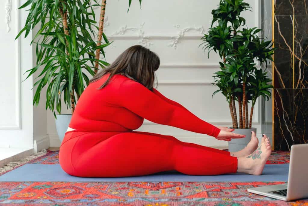 Loving myself in each moment, I ease into a seated forward bend on my yoga mat, surrounded by the gentle greenery of potted plants in a brightly lit room.