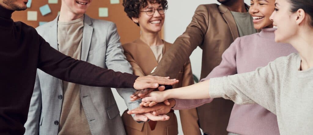 A diverse group of people stand in a circle, smiling and stacking their hands together in a gesture of teamwork.